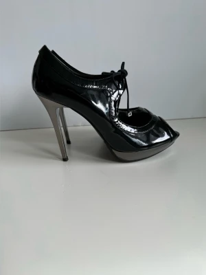 Svarta lackpumps från Karen Millen - storlek 40 - Snygga svarta pumps / högklackade skor i lack från Karen Millen i storlek 40, med hög smal klack i metall och öppen tå. Skorna har snörning framtill och en elegant, glansig finish som ger en cool och trendig look. 100% äkta läder. Välgjorda tidlösa kvalitetsskor. Mycket fint skick!