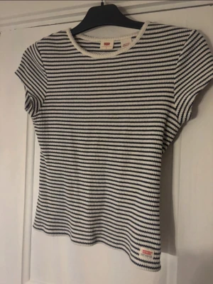 Randig topp från Levi's XS - Snygg ribbad topp från Levi's i vitt och marinblått med smala horisontella ränder. Modellen har korta ärmar, rund halsringning och liten logotyp nertill. Perfekt till jeans eller kjol för en avslappnad look. Oanvänd. Nypris 599:- 