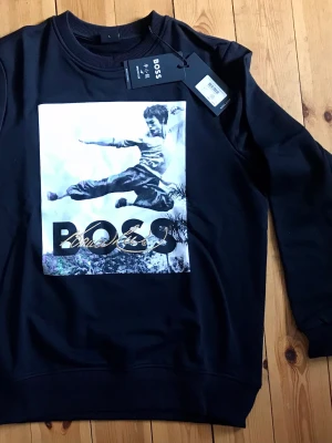 Svart BOSS x Bruce Lee sweatshirt - Cool svart sweatshirt/kofta från BOSS med stort tryck av Bruce Lee i action på framsidan och BOSS-logga. Tröjan har rund hals, ribbade muddar och är gjord i mjukt bomullsmaterial. Perfekt för dig som gillar streetwear och snygga prints.