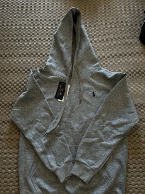 Grå hoodie från Polo Ralph Lauren - Snygg grå hoodie från Polo Ralph Lauren med klassisk huva och dragsnören. Broderad svart logga på bröstet och mjukt material som känns skönt mot huden. Perfekt för en avslappnad och stilren look.