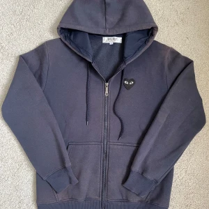 CDG Hoodie - Mörkblå hoodie från Comme des Garçons Play i storlek S. Tröjan är i fint skick förutom det att den är lite urtvättad på ärmarna och axlarna och därför har förlorat lite av sin färg, Syns mer på bild än vad det gör i verkligheten pågrund av det starka ljuset på bilderna. Nypris ligger på runt 2500kr,  men pågrund av defekterna så säljer jag därför hoodien för endast 799kr! Hör av er vid minsta frågor eller funderingar!