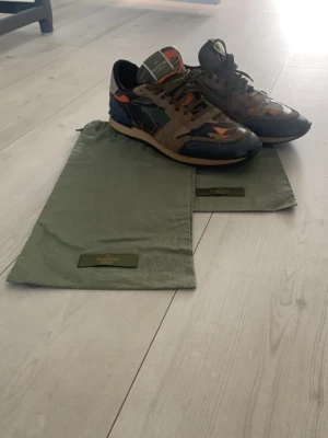 Valentino camo sneakers i mocka och läder - Snygga sneakers från Valentino med camo-mönster i grönt, orange och blått. Skorna har ovandel i mocka och läder, gröna skosnören och detaljer i flera färger. Kommer med original dustbags. Perfekta för dig som vill sticka ut med din stil.