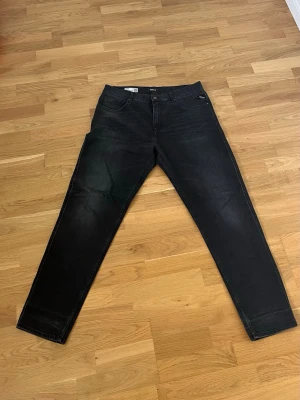 Svarta jeans från Replay - Säljer ett par svarta jeans från Replay med klassisk femficksdesign och raka ben. Jeansen har diskreta slitningar och är tillverkade i stretchigt denimtyg för extra komfort. Perfekta för en avslappnad och stilren look. 