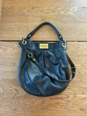 Marc by Marc Jacobs väska - Marc by Marc Jacobs bag with a hobo design in high quality material.   Perfect for everyday wear or a more dressed up occasion.   The bag comes with a shorter shoulder strap and a longer strap for the shoulder or crossbody.   The bag is in good condition with some small tears on the leather (mostly at the strap, see pictures) it’s not too noticeable when wearing it