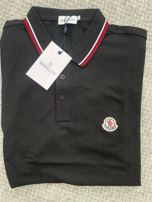 Svart pikétröja från Moncler - Klassisk svart pikétröja från Moncler med röd och vit rand på kragen. Tröjan har knappslå framtill och Moncler-logga broderad på bröstet. Perfekt för en stilren och sportig look.