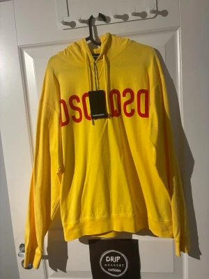 Gul hoodie från Dsquared2 - Gul hoodie från Dsquared2 med stor röd logga över bröstet. Klassisk modell med huva och dragsnören, samt ribbade muddar vid ärmslut och nederkant. Tillverkad i mjukt bomullsmaterial som känns skönt mot huden. Perfekt för en färgstark streetwear-look.