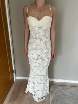 Vit lång spetsklänning med blommor - Elegant långklänning i vit spets med stora blommiga detaljer och smala axelband. Klänningen har figurnära passform och en lätt utsvängd nederdel. Perfekt för dig som vill ha en romantisk och stilren look.