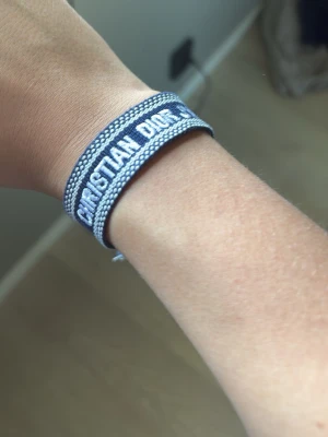 Blått armband från Christian Dior - Snyggt blått tygarmband från Christian Dior med vit broderad logga. Armbandet har justerbar knytning och dekorativa tofsar i ändarna. Perfekt accessoar för att lyfta din outfit med en lyxig touch.