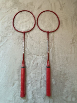 Badmintonset med två racketar - Säljer ett badmintonset med två röda racketar och två vita plastfjädrar. Racketarna har röda handtag och ram, samt vit snörning och badmintonstolpar. Perfekt för att spela badminton utomhus eller inomhus med kompisar.