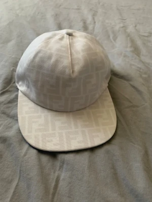 Vit keps från Fendi med FF-mönster - Snygg vit keps från Fendi med det klassiska FF-monogrammet ton-i-ton över hela kepsen. Kepsen har böjd skärm och är tillverkad i ett slitstarkt tygmaterial. Perfekt för dig som vill ha en stilren och exklusiv accessoar.