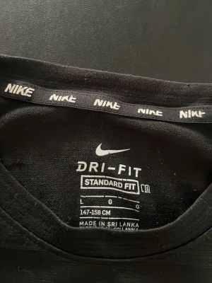 Svart Nike Dri-Fit t-shirt - Svart Nike Dri-Fit t-shirt med klassisk passform och korta ärmar. T-shirten har en liten vit Nike-logga på bröstet och är tillverkad i ett funktionsmaterial som andas. Perfekt för en sportig och avslappnad stil.