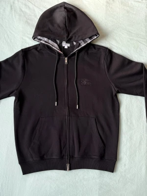 Svart hoodie från Burberry - Snygg svart hoodie från Burberry med dragkedja och huva. Klassisk design med diskret broderad logga på bröstet och randigt foder i huvan. Mjukt material och ribbade muddar. Perfekt för en chill och stilren look.