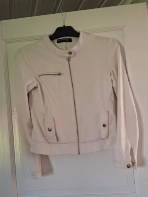 Vit  jacka med dragkedja - Snygg ljusbeige jacka från Outfitters Nation med rak passform, rund krage och dragkedja framtill. Jackan har en bröstficka med dragkedja och två fickor med lock och knapp. Perfekt för dig som gillar stilrena detaljer.