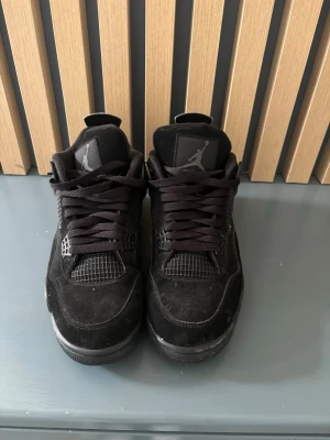 Svarta Air Jordan 4 Retro sneakers - Säljer ett par helsvarta Air Jordan 4 Retro sneakers med klassisk siluett, meshdetaljer och Jumpman-logga på plösen. Skorna har mockaliknande ovandel, svarta snören och gummisula. Perfekta för dig som gillar streetwear och ikoniska sneakers.