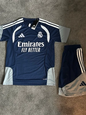 Real Madrid träningsset Adidas blå - Snyggt träningsset från Adidas för Real Madrid med tröja och shorts i marinblått och ljusgrått. Tröjan har klubbmärke, Adidas-logga och 'Emirates Fly Better' tryckt framtill. Setet är gjort i funktionsmaterial som andas och har klassiska vita ränder på axlar och shorts.