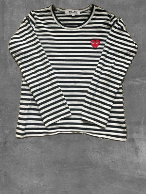 Randig långärmad T-shirt från Comme des Garçons - Svartvit randig långärmad T-shirt från Comme des Garçons PLAY med rund halsringning och ikoniskt rött hjärta på bröstet. Perfekt för en trendig och avslappnad look.