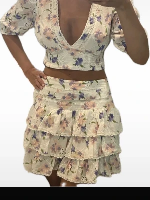 Blommig croppad blus och matchande kjol - Superfin croppad blus med v-ringning och puffärmar. Med en matchande kjol. Blusen är vit med blommigt mönster i lila och rosa toner, spetsdetaljer och broderade inslag. Perfekt för sommarens alla tillfällen!