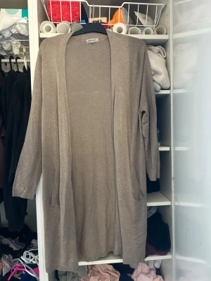 Beige lång kofta från Basic You - Lång, öppen beige kofta från Basic You i mjukt och tunt stickat material. Koftan har långa ärmar och två fickor framtill. Perfekt att slänga över en t-shirt eller topp för en avslappnad look.