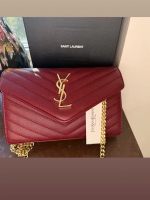 Vinröd YSL handväska med kedja - Snygg vinröd handväska från Saint Laurent med guldigt YSL-logo framtill. Väskan har quiltat mönster, guldfärgad kedjerem och magnetlås. Tillverkad i läder och har flera fack inuti för smart organisering. Äktighetsbevis saknas därav priset. Priset går att diskutera💞