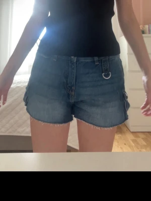 Bershka jeans shorts - Säljer en blå jeans shorts i klassisk denim med rå kant nertill och två stora fickor med knappar på sidan. Shortsen har normal passform och är perfekt för dig som gillar en avslappnad men ändå trendig stil.