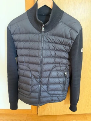 Svart pufferjacka från Moncler - Snygg svart pufferjacka från Moncler med quiltad front och rygg i glansigt material samt stickade ärmar och krage. Jackan har dragkedja framtill, två fickor med dragkedja och Moncler-logga på ärmen. Perfekt för kalla vinterdagar.