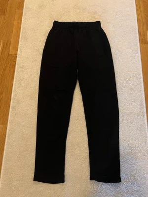 Nike mjukisbyxor - Svarta mjukisbyxor från Nike med rak passform och resår i midjan. Byxorna har en enkel design och en bakficka. Perfekta för en avslappnad stil och tillverkade i mjukt material för extra komfort.