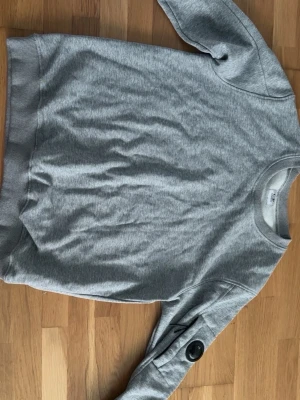 Grå sweatshirt från C.P. Company - Snygg grå sweatshirt från C.P. Company med klassisk rund hals och ribbade muddar. Tröjan har en ikonisk linsdetalj på ärmen som ger en unik touch. Perfekt för en avslappnad och stilren look.