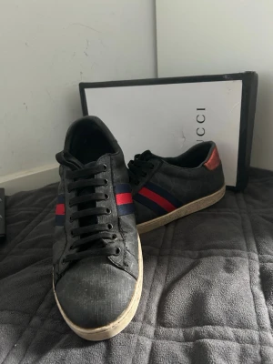 Gg skor  - Snygga svarta sneakers med klassiska röda och blå ränder på sidan. Skorna har lågt skaft och en vit sula med unikt mönster undertill. Materialet är canvas med läderdetaljer och en röd hälflik. Perfekta för dig som gillar exklusiv streetstyle. Skador i sulan och är välanvända storlek 43
