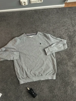 Grå sweatshirt från Ralph Lauren  - Snygg grå sweatshirt från Ralph Lauren - U.S Polo Assasin med klassisk rund hals och broderad logga på bröstet. Tröjan har ribbade muddar vid ärmslut och nederkant. Perfekt för en avslappnad och stilren look på en utekväll. Tröjan är relativt tun och sval under sommarkvällar, Storlek M. 