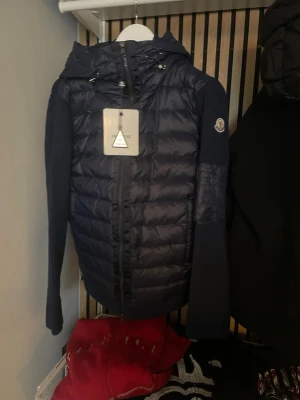 Moncler tricot cardigan - Snygg mörkblå pufferjacka från Moncler med quiltad front, ribbstickade ärmar och huva. Jackan har dragkedja framtill och Moncler-logga på ärmen. Perfekt för kalla dagar när du vill hålla stilen och värmen.