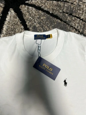 Vit t-shirt från Polo Ralph Lauren - Vit t-shirt från Polo Ralph Lauren med klassisk rund hals och liten broderad logga på bröstet. Tidlös design som passar till allt och är tillverkad i mjuk bomull för maximal komfort.