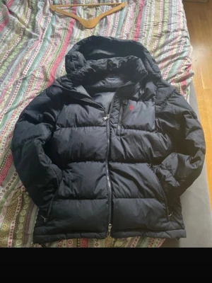 Svart pufferjacka från Polo Ralph Lauren - Säljer en svart pufferjacka från Polo Ralph Lauren med röd broderad logga på bröstet. Jackan har huva, dragkedja framtill och två sidofickor. Perfekt för kalla dagar och riktigt skön att bära. Klassisk och stilren design som funkar till det mesta. De ända är att handspänne grejen är sönder se bild 6 men de är ingen big deal och går att fixa annars är de typ nyskick