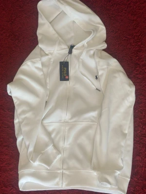 Vit hoodie från Polo Ralph Lauren - Snygg vit hoodie från Polo Ralph Lauren med dragkedja framtill och klassisk logga broderad på bröstet. Hoodien har huva med dragsko och ribbade muddar vid ärmslut och nederkant. Perfekt för en clean och stilren look.
