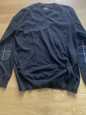 Svart tröja med rutiga armbågslappar - Klassisk brun tröja från Barbour med V-ringning och rutiga armbågslappar i mörka färger. Tröjan har långa ärmar och ribbade muddar vid ärmslut och nederkant. 