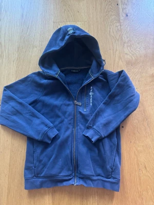 Marinblå hoodie från Sail Racing - Snygg marinblå hoodie från Sail Racing med dragkedja framtill och broderad logga på bröstet. Hoodien har en mysig fleeceinsida, två fickor och en huva med randig detalj. Perfekt för chill dagar eller när du vill ha en sportig look.
