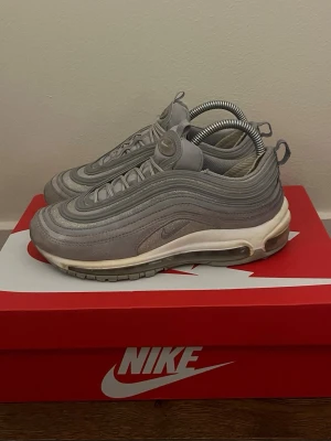 Nike Air Max 97 grå sneakers - Nike Air Max 97 sneakers i helgrå färg med ikoniska vågformade paneler och synlig Air Max-sula. Ovandelen är i textil och syntet med reflekterande detaljer och klassisk Nike-logga på plösen. Skorna har en sportig och futuristisk look.