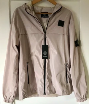 Beige vindjacka från Stone Island - Snygg beige vindjacka från Stone Island med huva och hel dragkedja framtill. Jackan har svarta detaljer, två fickor med dragkedja och klassisk logotyp på ärmen. Perfekt för en stilren och sportig look. Materialet är lätt och vindtätt.