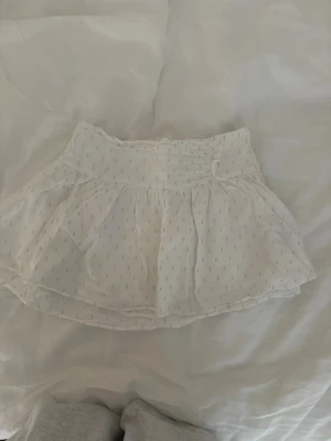 Vit kortkjol med diskret mönster - Söt vit kortkjol med lätt volang och diskret prickigt mönster. Kjolen har bred resår i midjan och är gjord i ett tunt, luftigt material som ger en flowy känsla. Perfekt för en fräsch och somrig look. Den är från Ginas barnavdelning i storlek 158/164 men skulle säga att den är som en XS