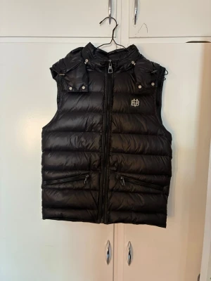Svart dunväst från bvaldi i nyskick - Snygg svart dunväst från bvaldi med hög krage, två dragkedjeförsedda fickor och logotyp på bröstet. Västen är slimfit och har quiltad design och detaljer med tryckknappar vid axlarna. Perfekt för lager-på-lager och streetstyle.
