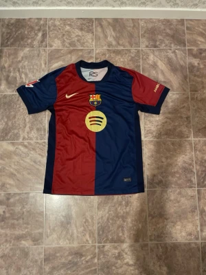 FC Barcelona matchtröja Lamine Yamal - Säljer en FC Barcelona matchtröja från Nike med Lamine Yamal och nummer 19 på ryggen. Tröjan är röd och marinblå med klubbmärke, Nike-logga och Spotify som sponsor framtill. Korta ärmar och tillverkad i lätt, ventilerande material.