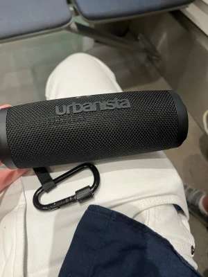 Urbanista trådlös högtalare svart - Snygg svart Urbanista trådlös högtalare med robust cylinderformad design och textilöverdrag. Utrustad med tydliga knappar för volym, play/pause och Bluetooth samt en praktisk karbinhake för enkel upphängning. Perfekt för musik på språng.