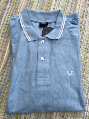Ljusblå pikétröja från Fred Perry - Snygg ljusblå pikétröja från Fred Perry med klassisk krage och vita ränder. Tröjan har knappar framtill och den ikoniska Fred Perry-loggan broderad på bröstet. Perfekt för en stilren och avslappnad look.