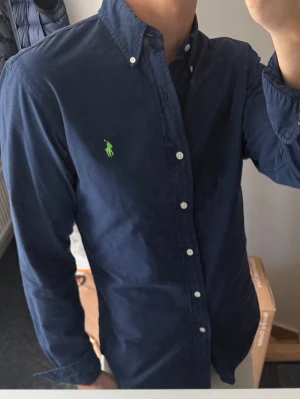 Mörkblå skjorta från Ralph Lauren - Klassisk mörkblå skjorta från Ralph Lauren med button-down krage och vita knappar. Skjortan har lång ärm och den ikoniska gröna broderade loggan på bröstet. Perfekt för en stilren look och tillverkad i bomull.