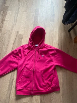 Rosa zip-hoodie från Adidas - Snygg rosa hoodie från Adidas med dragkedja framtill och klassisk huva med dragsko. Tröjan har två stora fickor på magen och Adidas logga broderad på bröstet. Tillverkad i mjukt sweatshirtmaterial som är skönt att ha på sig.