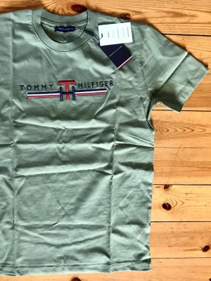 Grön Tommy Hilfiger t-shirt - Snygg oliv  t-shirt från Tommy Hilfiger med broderad logga och ränder i rött, vitt och blått på bröstet. Klassisk rund hals och korta ärmar. Perfekt för en stilren och avslappnad look.