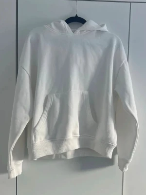 Vit hoodie med känguruficka - Säljer en stilren vit hoodie med huva och stor känguruficka framtill. Hoodien har ribbade muddar vid ärmslut och nederkant. Perfekt för en avslappnad och clean look. Materialet känns mjukt och bekvämt mot huden.