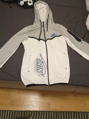 Nike vit och grå vindjacka med huva - Nike vindjacka i vitt och ljusgrått med svart dragkedja och detaljer. Stor blå Nike-logga på framsidan och mindre logga på bröstet. Jackan har huva med dragsko, långa ärmar och två fickor framtill. Perfekt för blåsiga dagar.