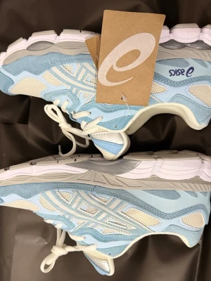 Asics sneakers i blått och vitt - Snygga sneakers från Asics i ljusblått och vitt med meshpaneler och detaljer i mocka. Skorna har vita snören, tjock sula och Asics-logga på sidan och hälen. Perfekta för en sportig och trendig look.
