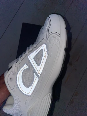 Vita CD B30 sneakers med reflexdetaljer - Säljer ett par vita B30 sneakers med tydliga reflexdetaljer och stora loggor på sidorna. Skorna har en sportig design med mesh och läderpaneler, samt tjock sula för extra komfort. Perfekta för dig som gillar exklusiva och trendiga sneakers. Testade en gång! 