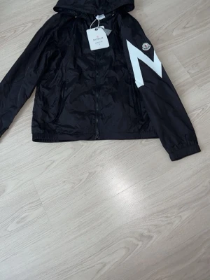 Svart vindjacka från Moncler - svart vindjacka från Moncler med vit blixtdetalj på sidan och klassisk Moncler-logga. Omvänd storlek S 